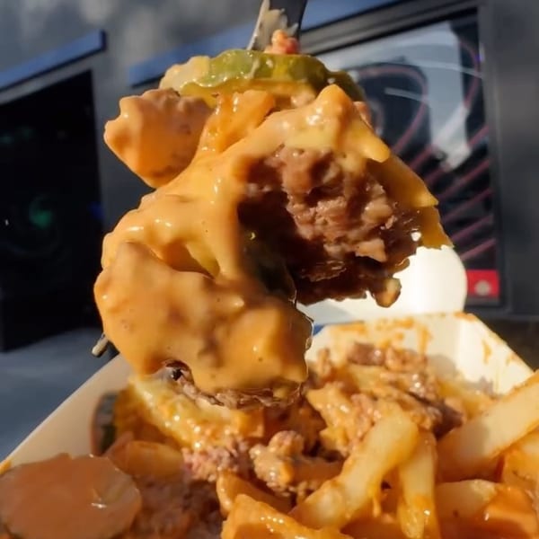 A fork lifts a cheesy burger bite with pickles from a container of loaded fries.