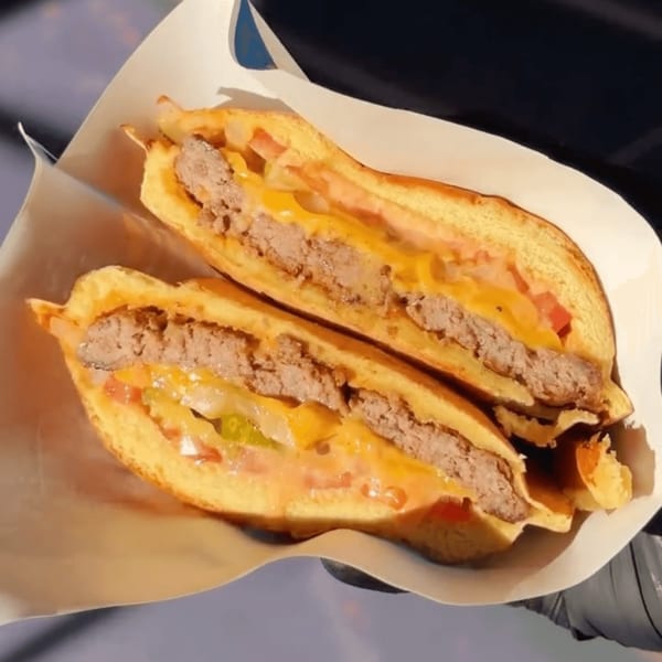 A halved Astroid burger with melted cheese, beef patties, tomatoes, and pickles, wrapped in paper.