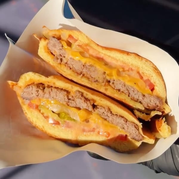 Two halves of a burger with cheese, tomatoes, and pickles, served in a paper wrapper.